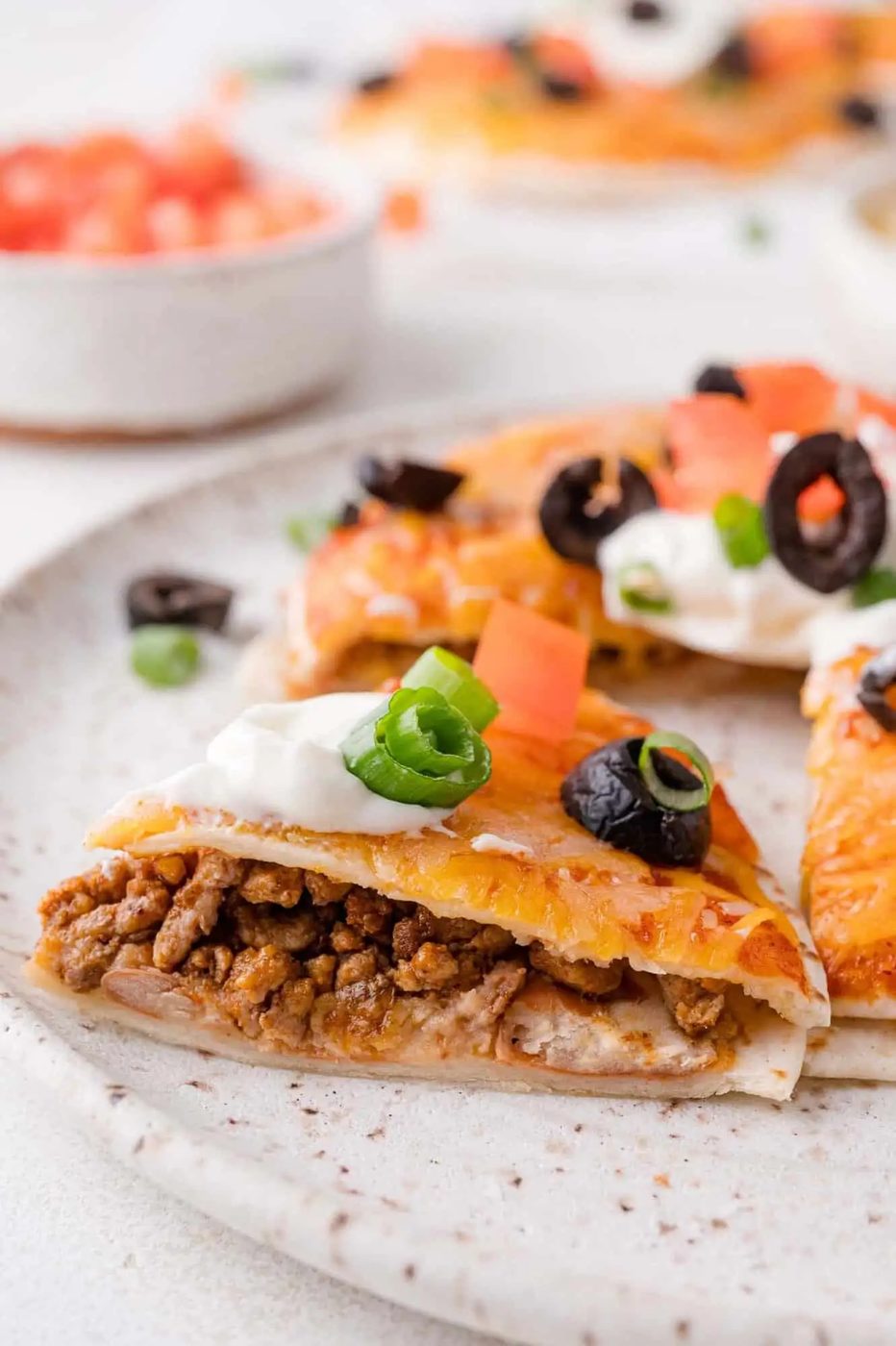 Close-up of a slice of a beef Mexican pizza topped with sour cream, diced tomatoes, sliced black olives, and green onions on a plate.
