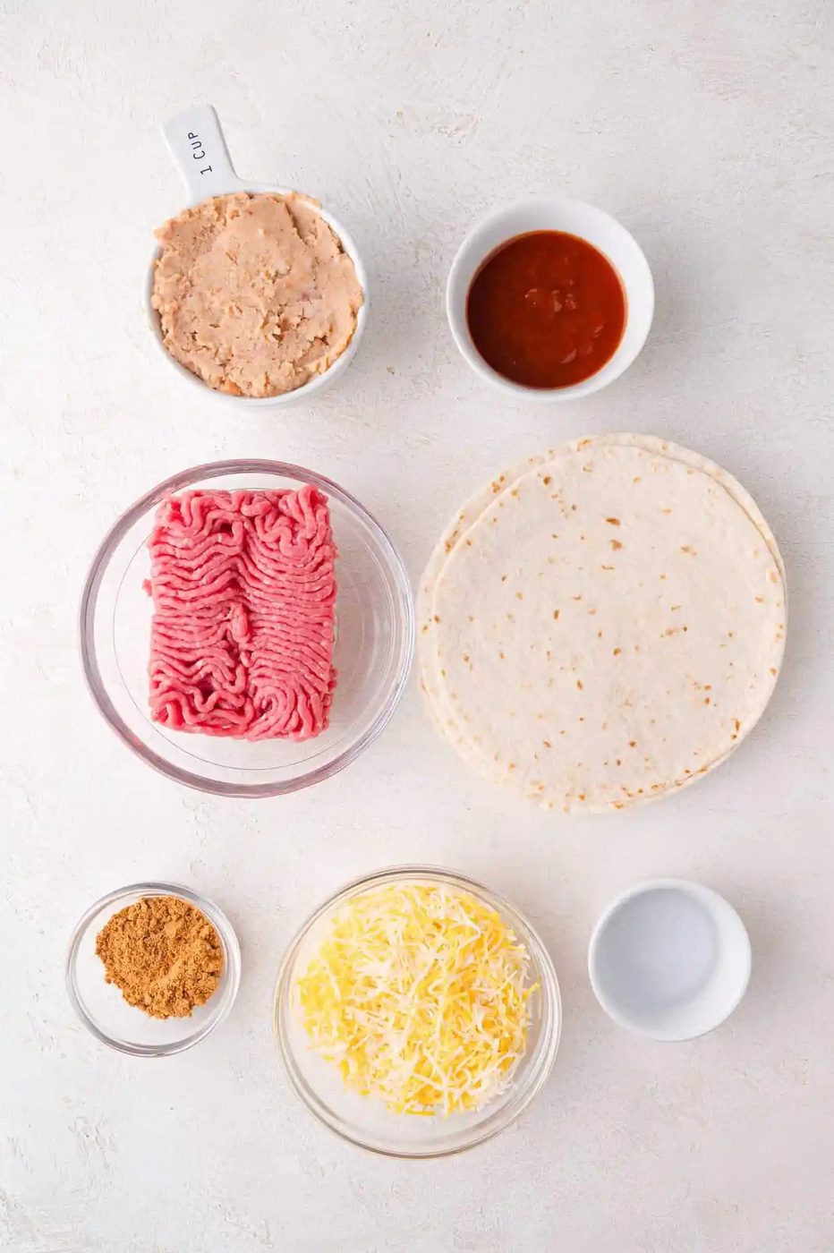 Ingredients arranged on a white surface include raw ground meat, tortillas, shredded cheese, refried beans, salsa, taco seasoning, and an empty measuring cup.