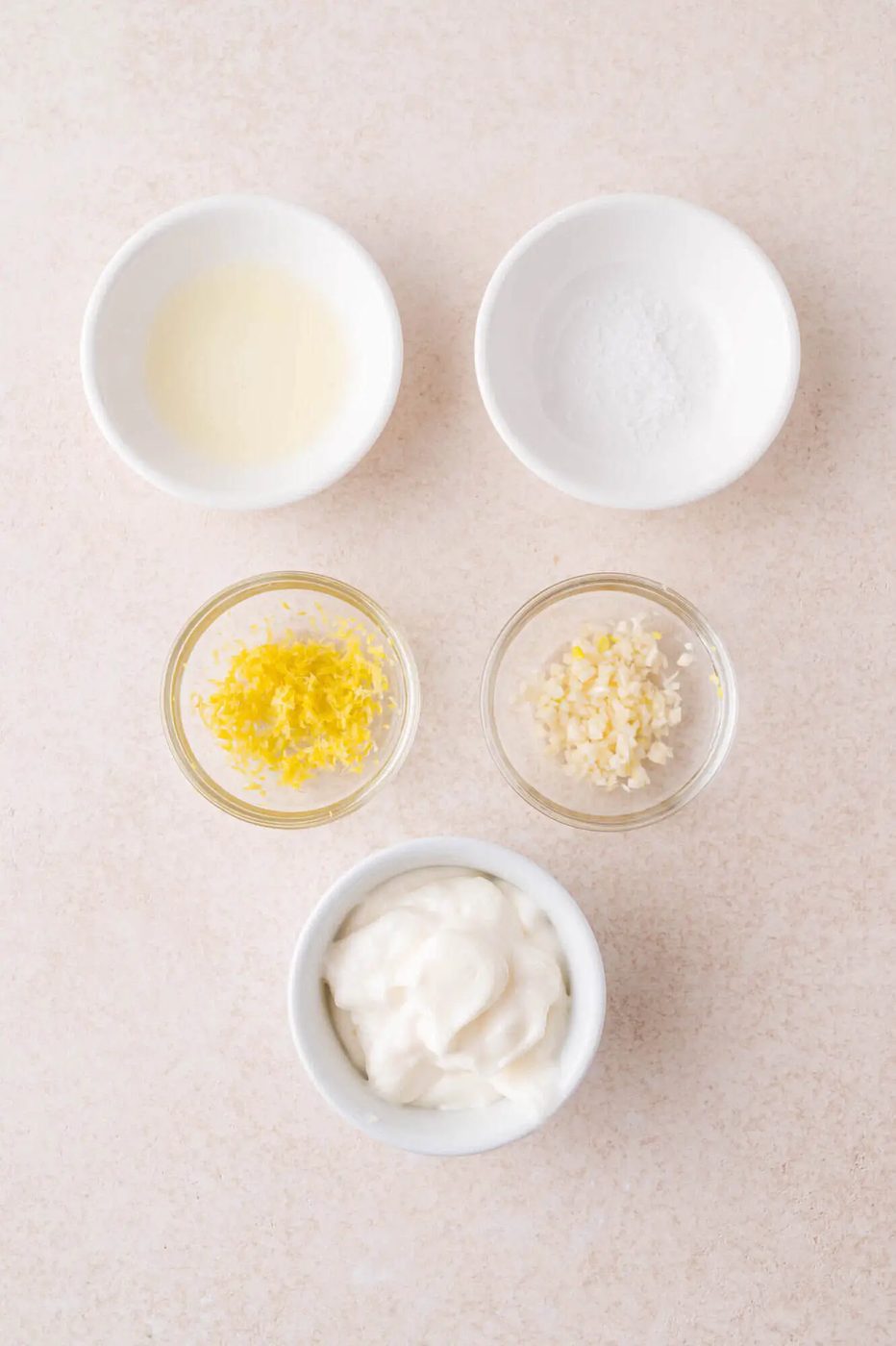Five small bowls containing lemon juice, salt, lemon zest, minced garlic, and mayonnaise arranged on a light surface.