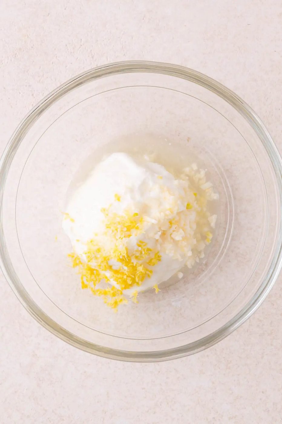 A glass bowl containing yogurt, minced garlic, lemon zest, and lemon juice on a light-colored surface.