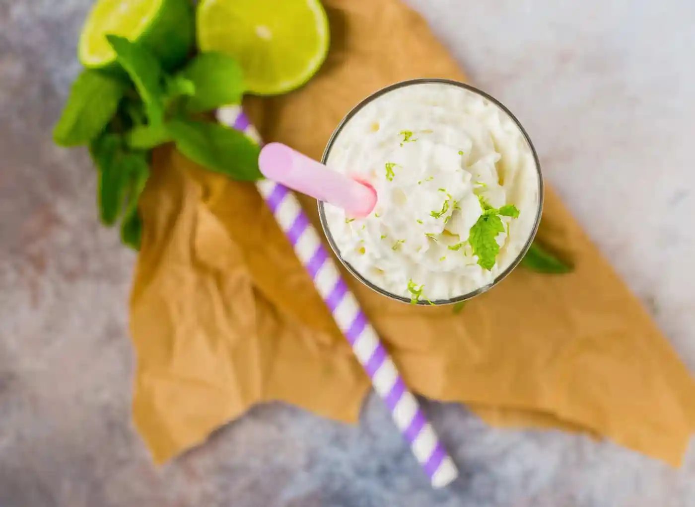 A glass of whipped cream-topped milkshake with lime zest and mint, a pink straw, two striped straws, lime slices, and mint leaves on brown paper.