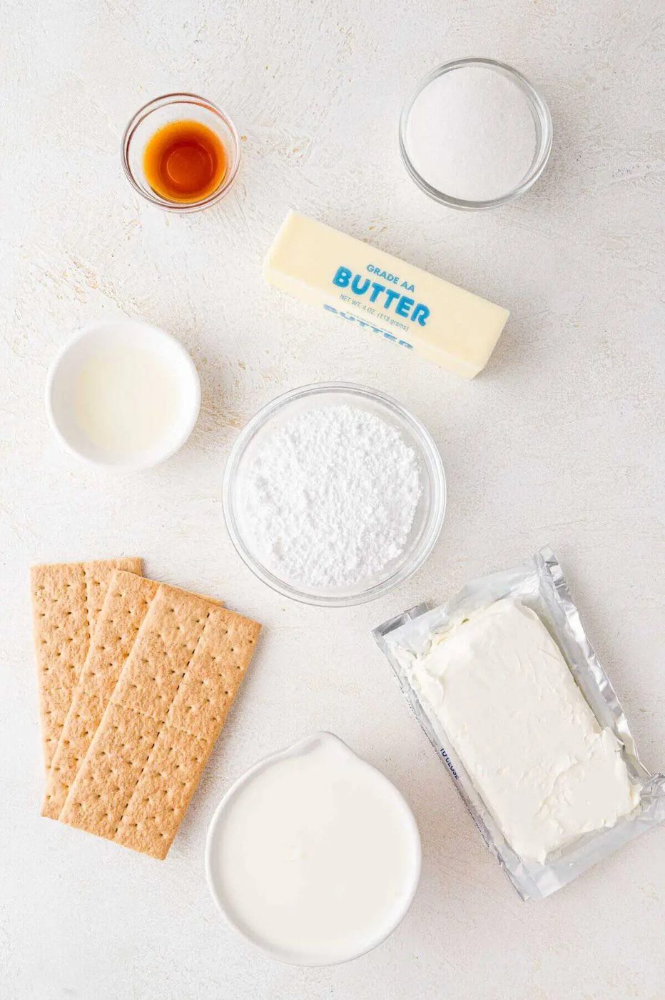 Ingredients for a recipe arranged on a table: vanilla extract, sugar, butter, milk, powdered sugar, cream cheese, and graham crackers.