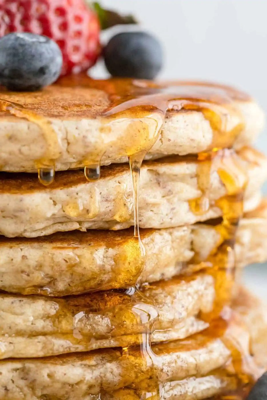 Stack of pancakes with syrup dripping down, topped with blueberries and a sliced strawberry.