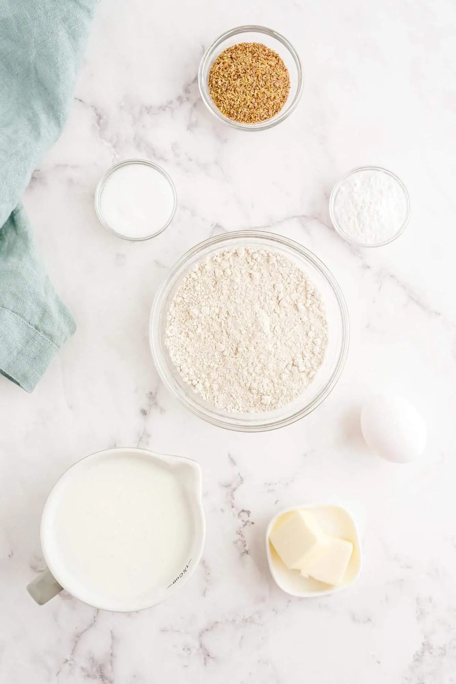 Ingredients on a marble countertop include milk, butter, an egg, flour, sugar, baking powder, and brown sugar in small bowls and a pitcher. A folded cloth is at the top left corner.