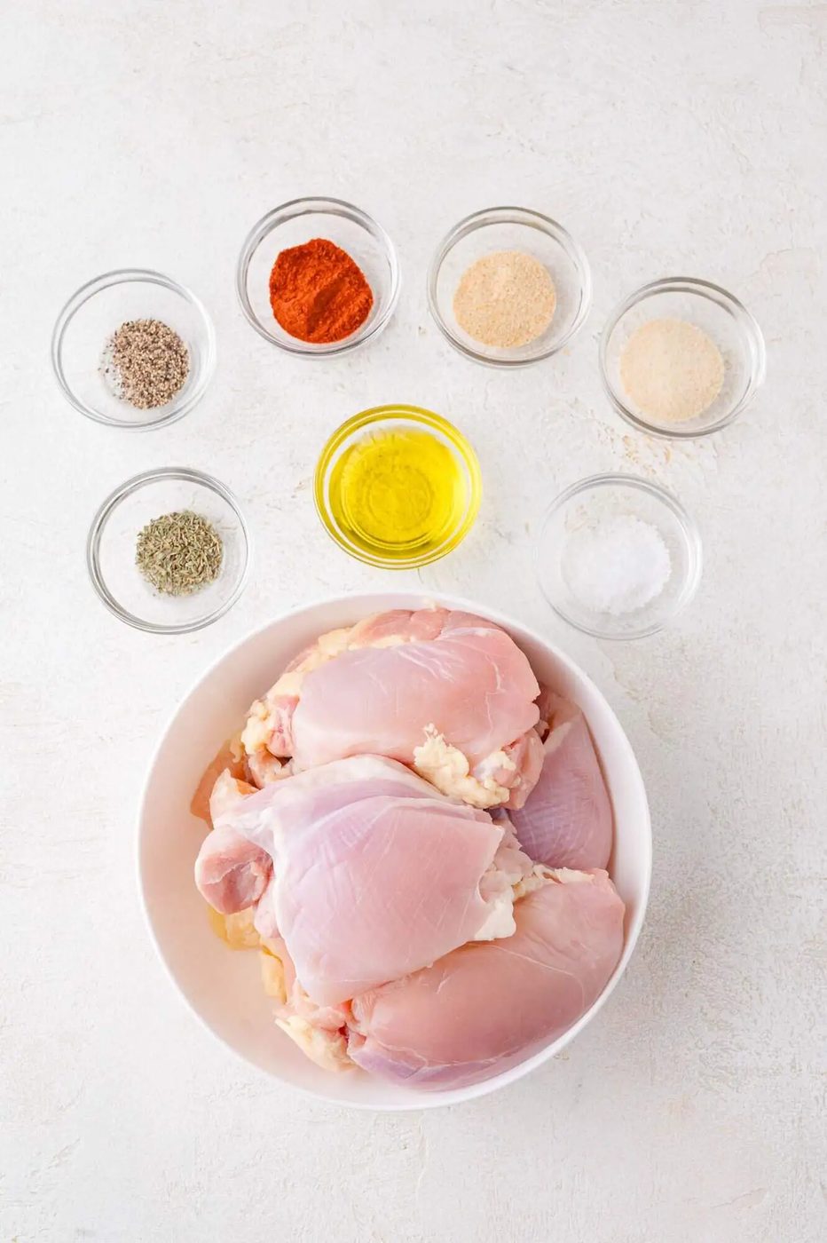Raw chicken thighs in a white bowl, surrounded by small glass bowls containing oil, salt, black pepper, paprika, garlic powder, onion powder, and dried herbs on a white surface.