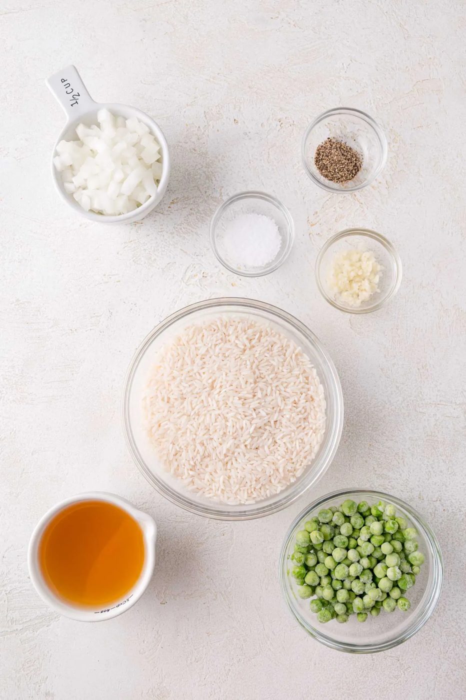Bowls containing chopped onions, salt, pepper, minced garlic, white rice, frozen peas, and a measuring cup of vegetable broth arranged on a white surface.