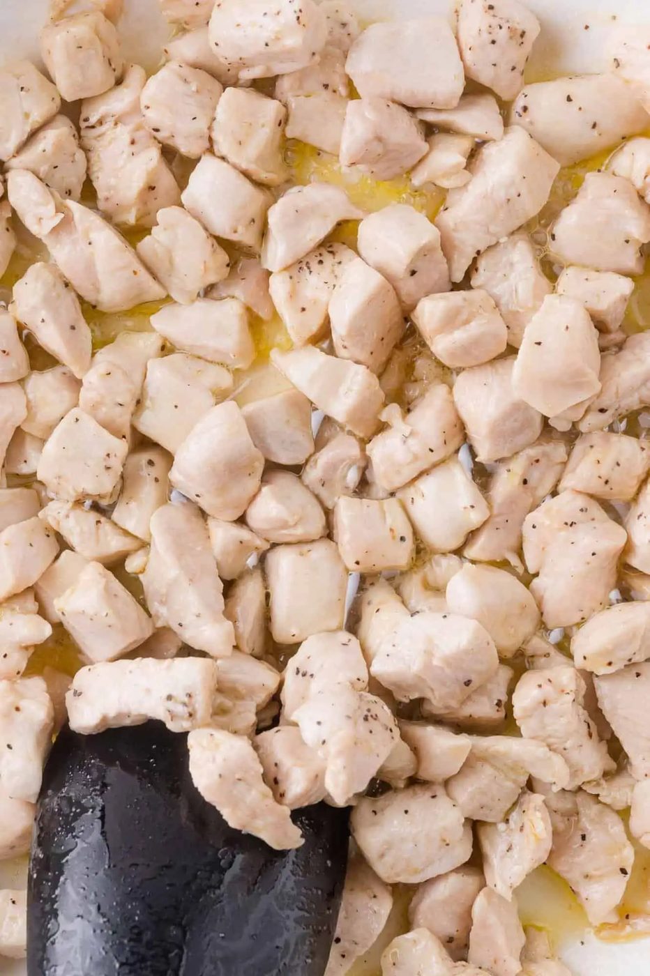 Cubed pieces of chicken breast are being cooked in a pan, with a black spatula stirring the lightly browned and seasoned meat.