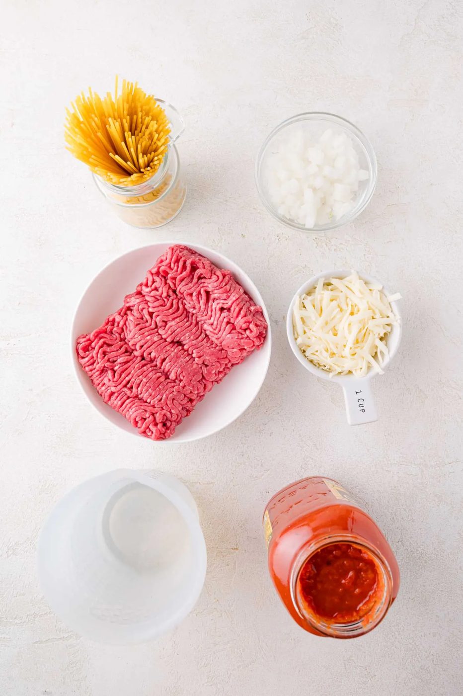 Uncooked spaghetti, chopped onions, shredded cheese, ground beef, marinara sauce, and a measuring cup of water arranged on a white surface.