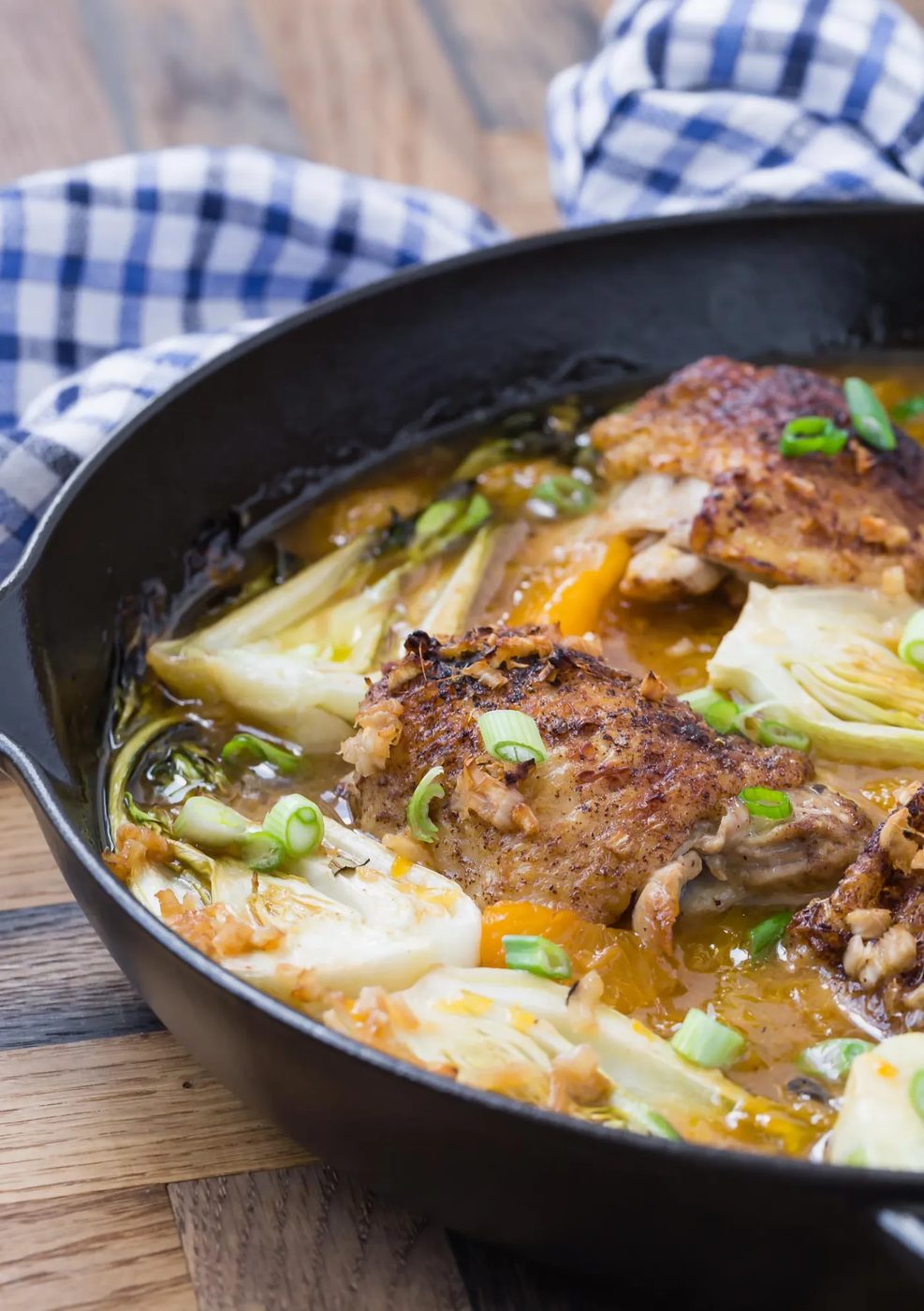 Black skillet with orange chicken thighs and bok choy, on a wooden surface.