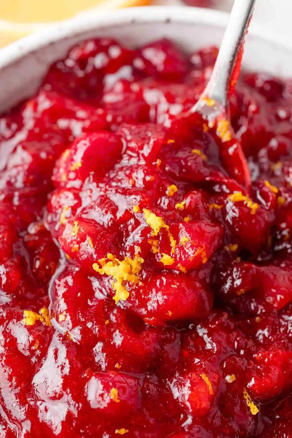 Close-up of a bowl of cranberry sauce with visible orange zest on top, and a spoon partially submerged in the sauce.