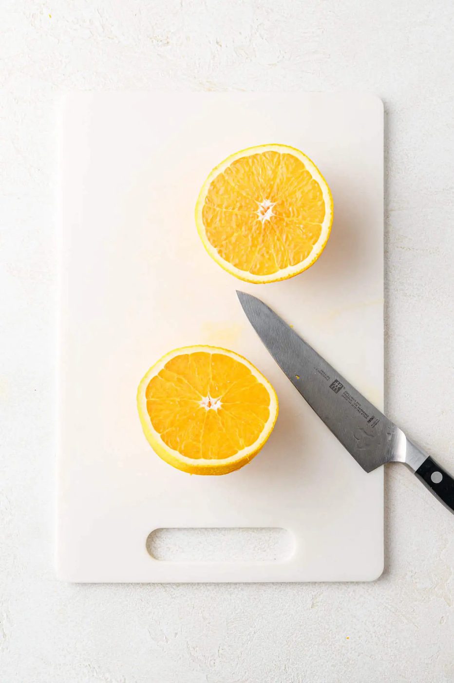 Two orange halves and a knife on a white cutting board.