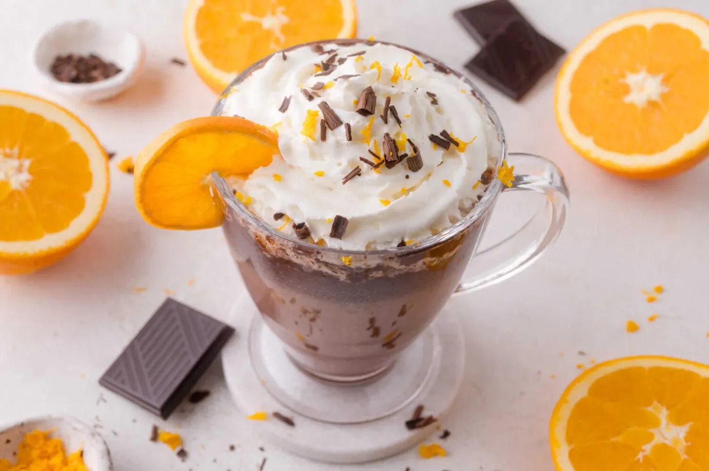 A glass mug of hot chocolate topped with whipped cream, chocolate shavings, and orange zest, garnished with an orange slice, surrounded by orange halves and chocolate pieces.