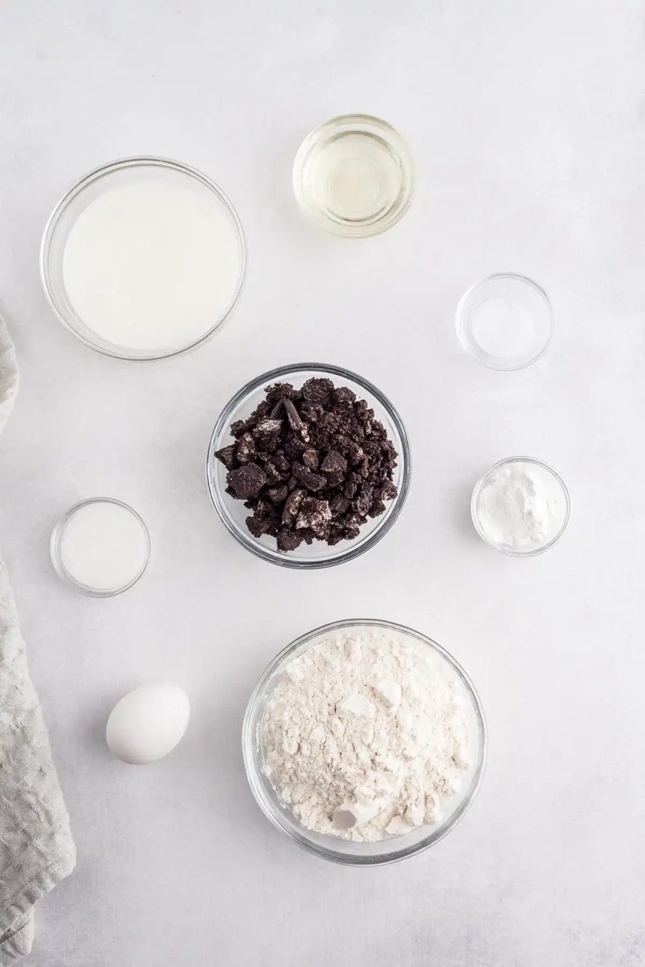 Baking ingredients on a table: bowl of milk, crushed cookies, flour, egg, sugar, baking powder, and oil.