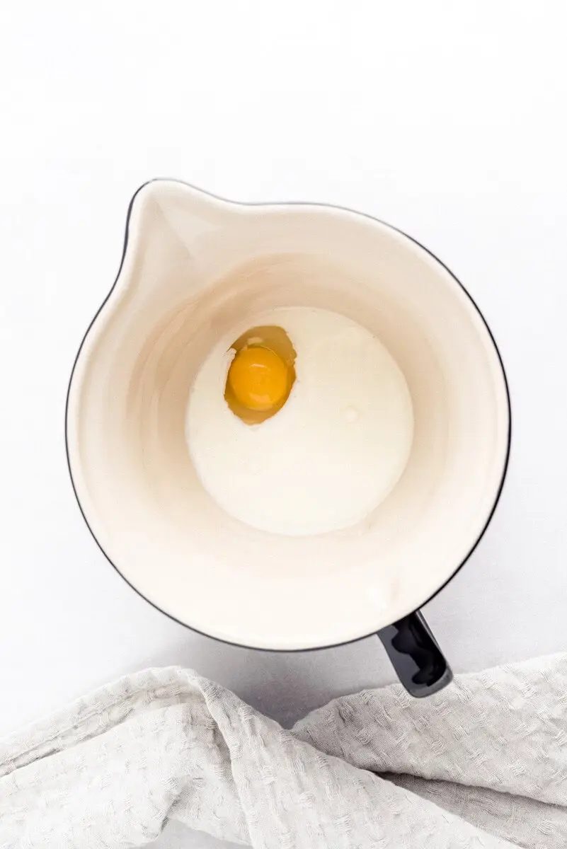 A single egg yolk and milk in a ceramic mixing bowl, placed on a white towel with a subtle pattern.