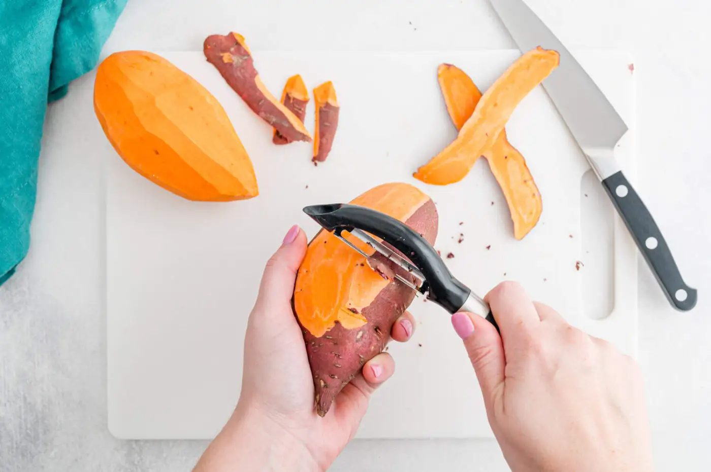 A person peels a sweet potato with a vegetable peeler on a white cutting board next to a knife, peels, and a partially peeled sweet potato.