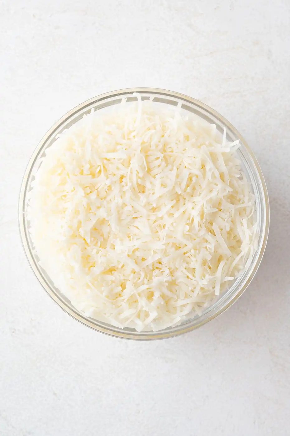 A clear glass bowl filled with shredded Parmesan cheese sits on a white surface.