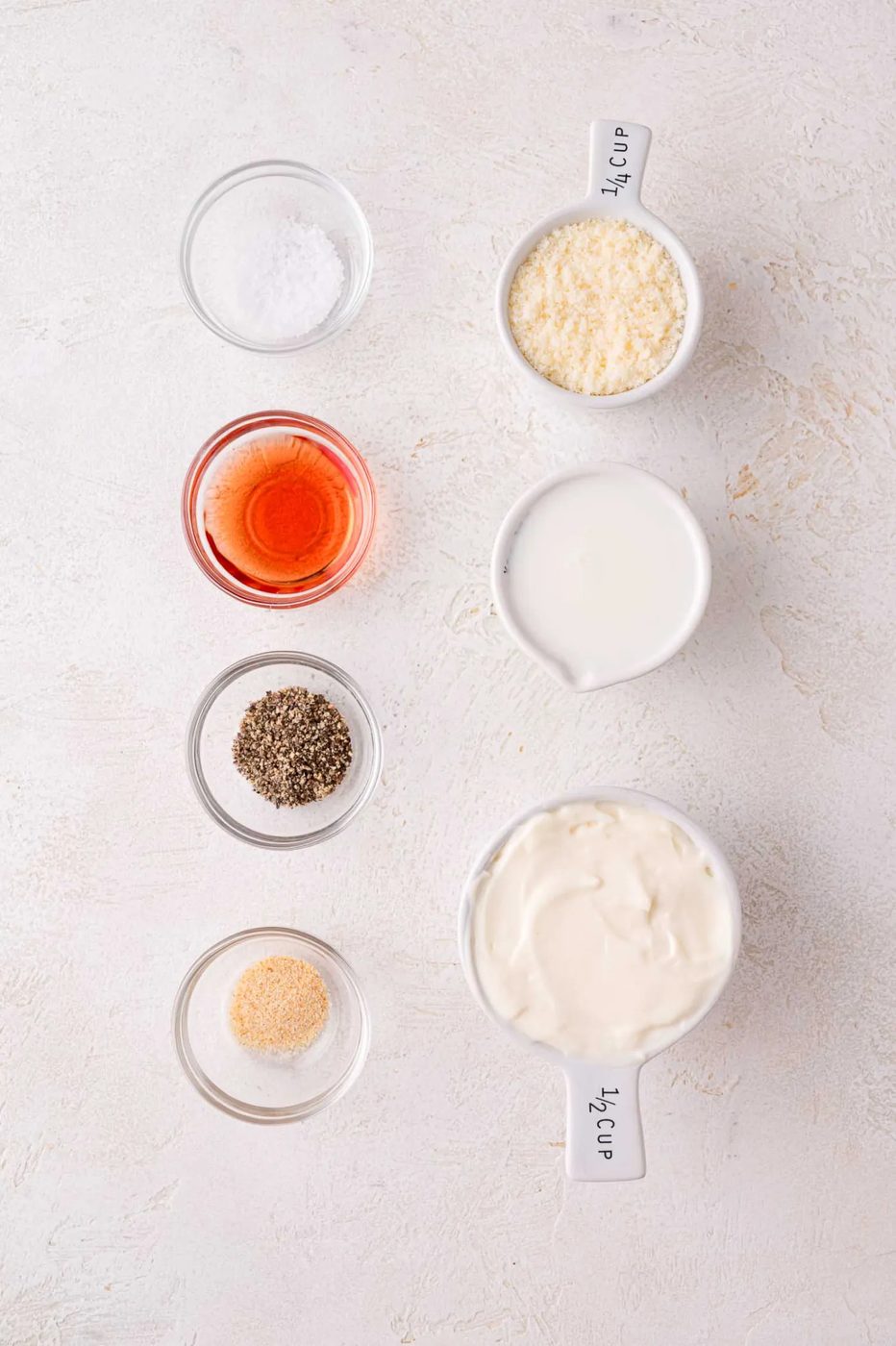 Six small bowls and cups containing salt, grated Parmesan, red vinegar, milk, black pepper, garlic powder, and mayonnaise arranged on a light surface.