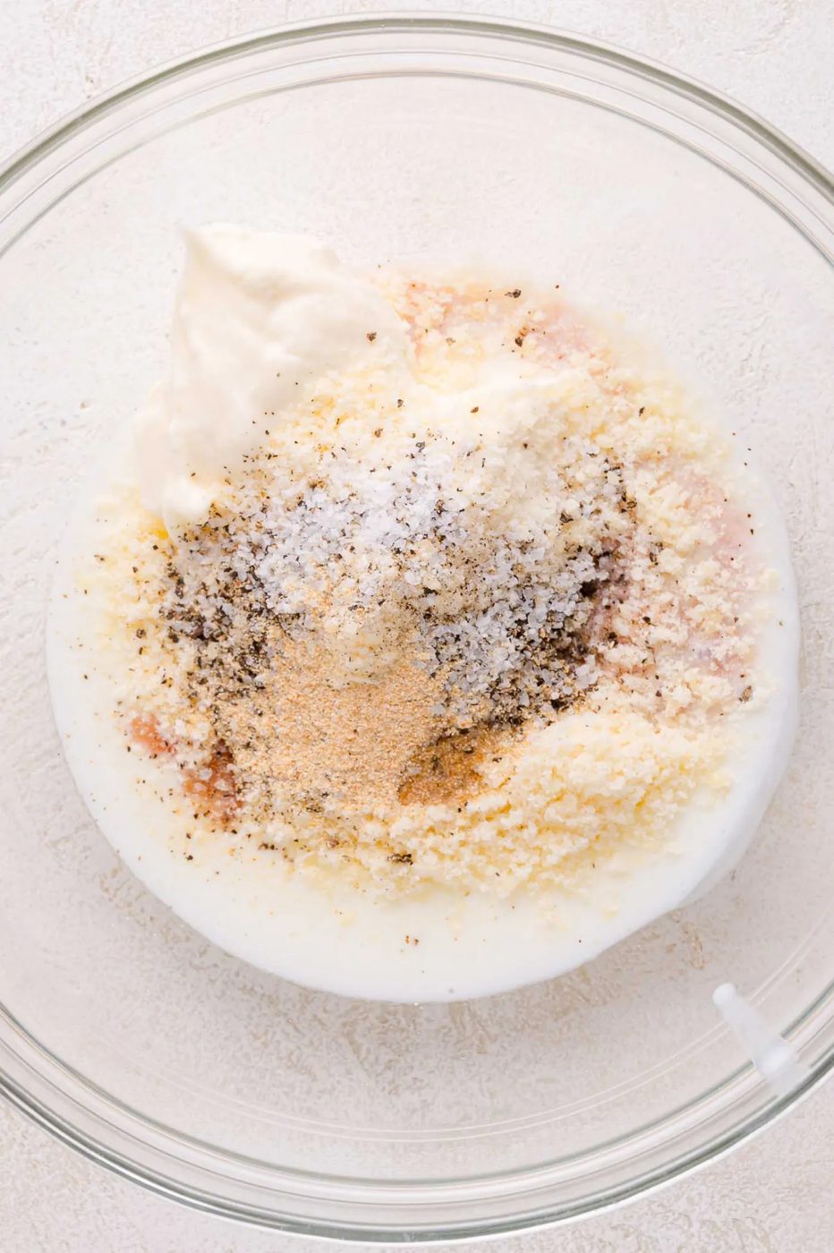 A glass bowl containing mayonnaise, grated cheese, black pepper, salt, garlic powder, and other seasonings, viewed from above.