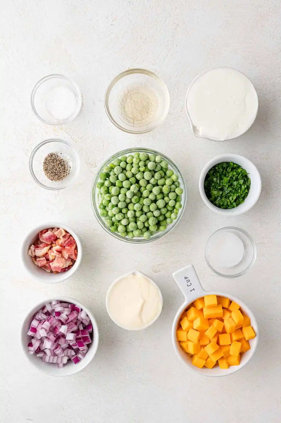 Various ingredients in bowls on a white surface, including frozen peas, diced cheddar, chopped red onion, mayonnaise, cooked bacon, sour cream, parsely, vinegar, salt, pepper, and sugar.