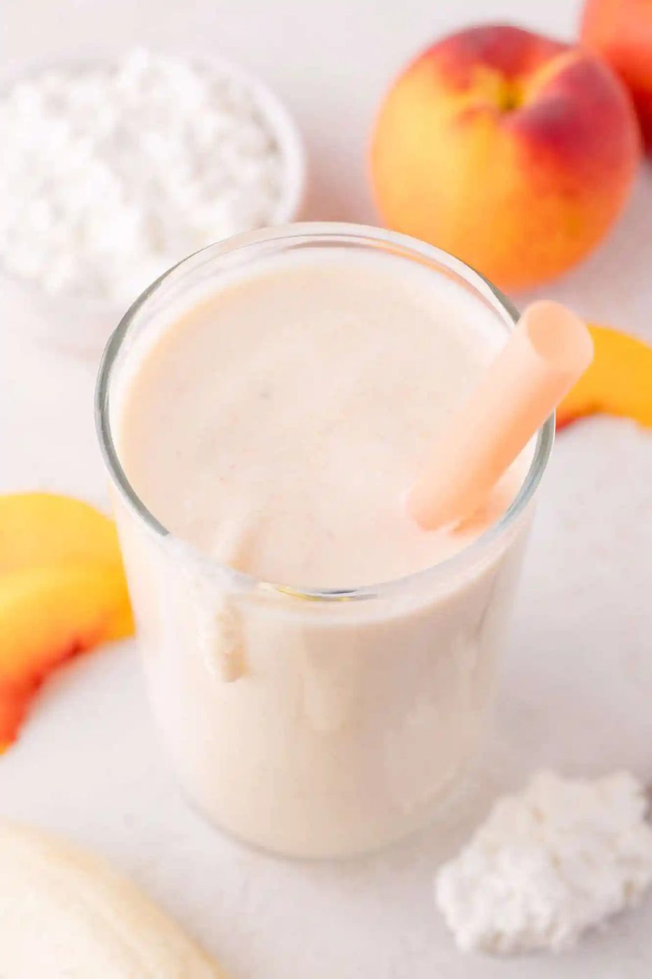 A glass of creamy smoothie with a straw, surrounded by peach slices, a whole peach, cottage cheese, and a banana on a white surface.