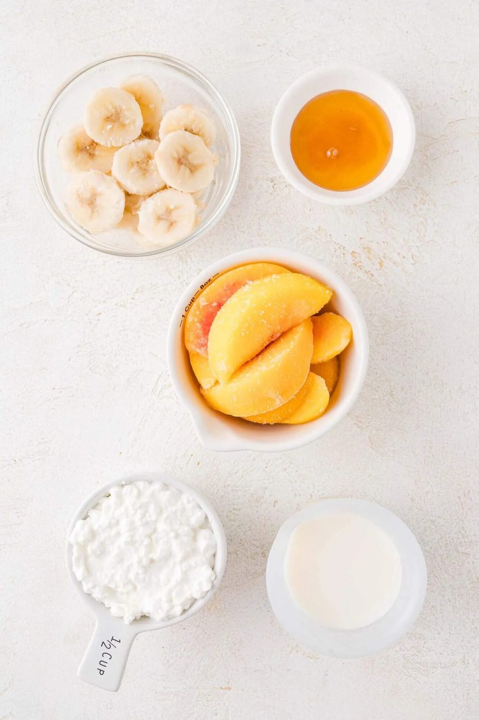 Top-down view of sliced bananas, peach slices, honey, cottage cheese, and milk in separate bowls on a white surface.