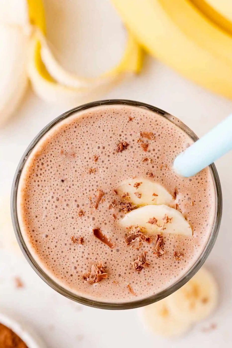 A glass of chocolate banana milkshake with a blue straw, topped with chocolate shavings and banana slices.