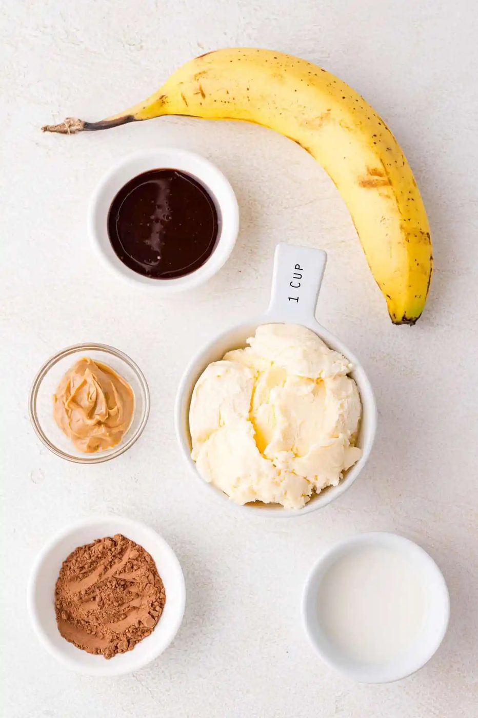 Banana, ice cream, cocoa powder, peanut butter, chocolate sauce, and milk arranged on a white surface.