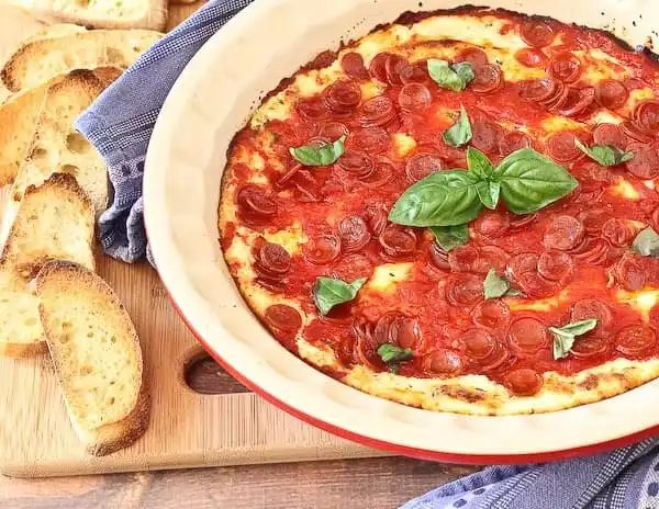 Front view of baked pizza dip on wooden cutting board with blue towel.