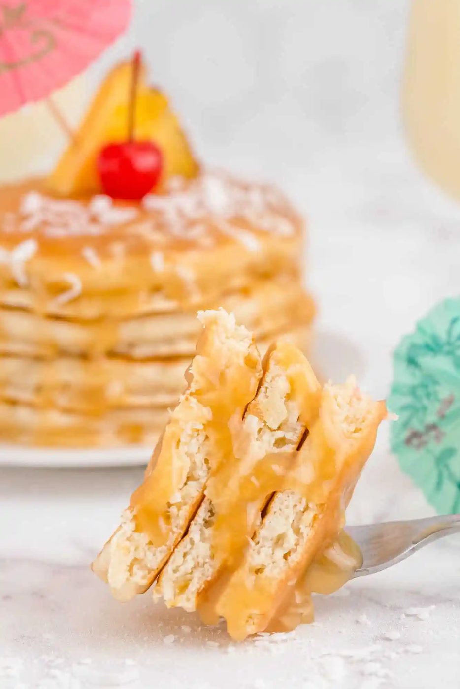 A fork holds a bite of pancakes drizzled with syrup, with a stack of decorated pancakes and cocktail umbrellas in the background.