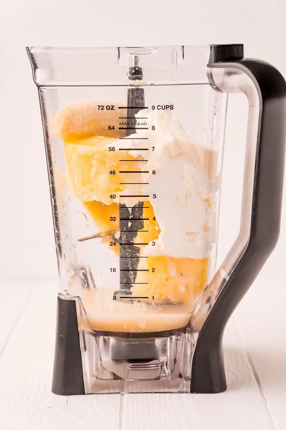 A blender pitcher containing scoops of yellow pineapple and white yogurt, with measurements marked on the side, sits on a white surface.