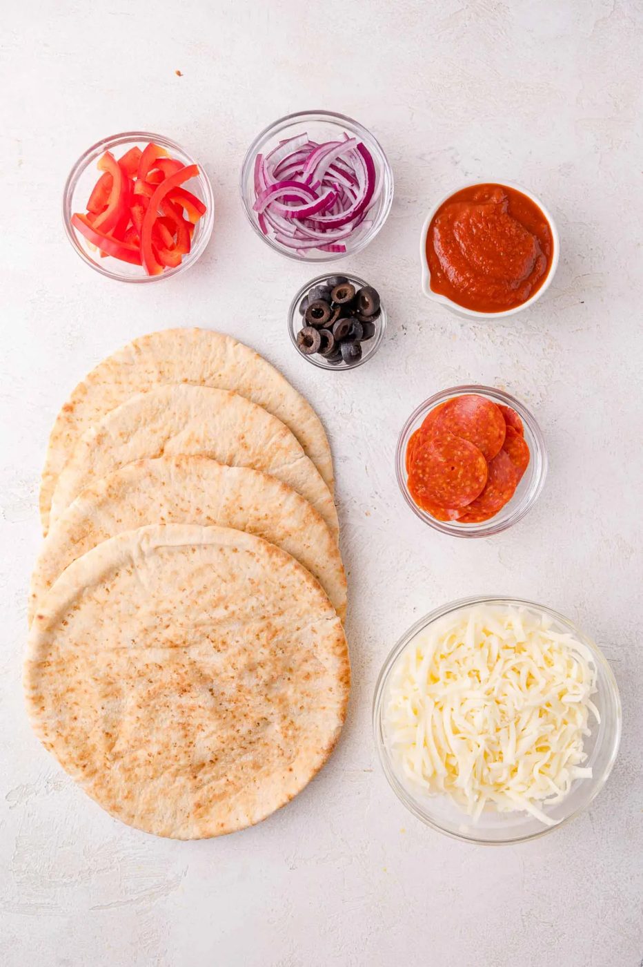 Four pieces of pita bread with bowls of shredded mozzarella, pepperoni slices, pizza sauce, black olives, red onion, and sliced red bell pepper on a white surface.
