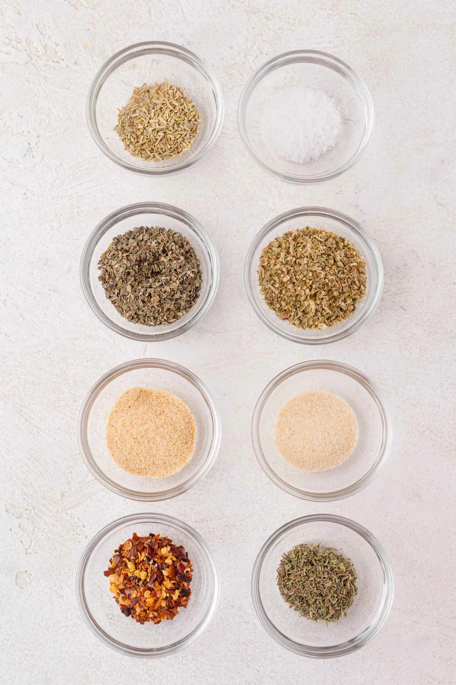 Eight small glass bowls filled with different dried herbs and spices, arranged in two vertical columns on a light surface.