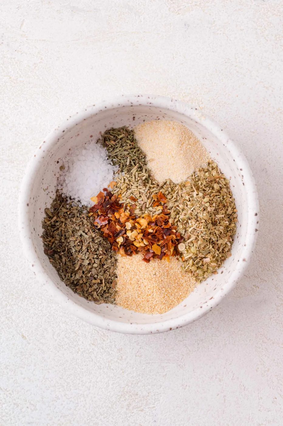 A white bowl containing separate piles of dried herbs, crushed red pepper flakes, salt, garlic powder, and onion powder on a light surface.