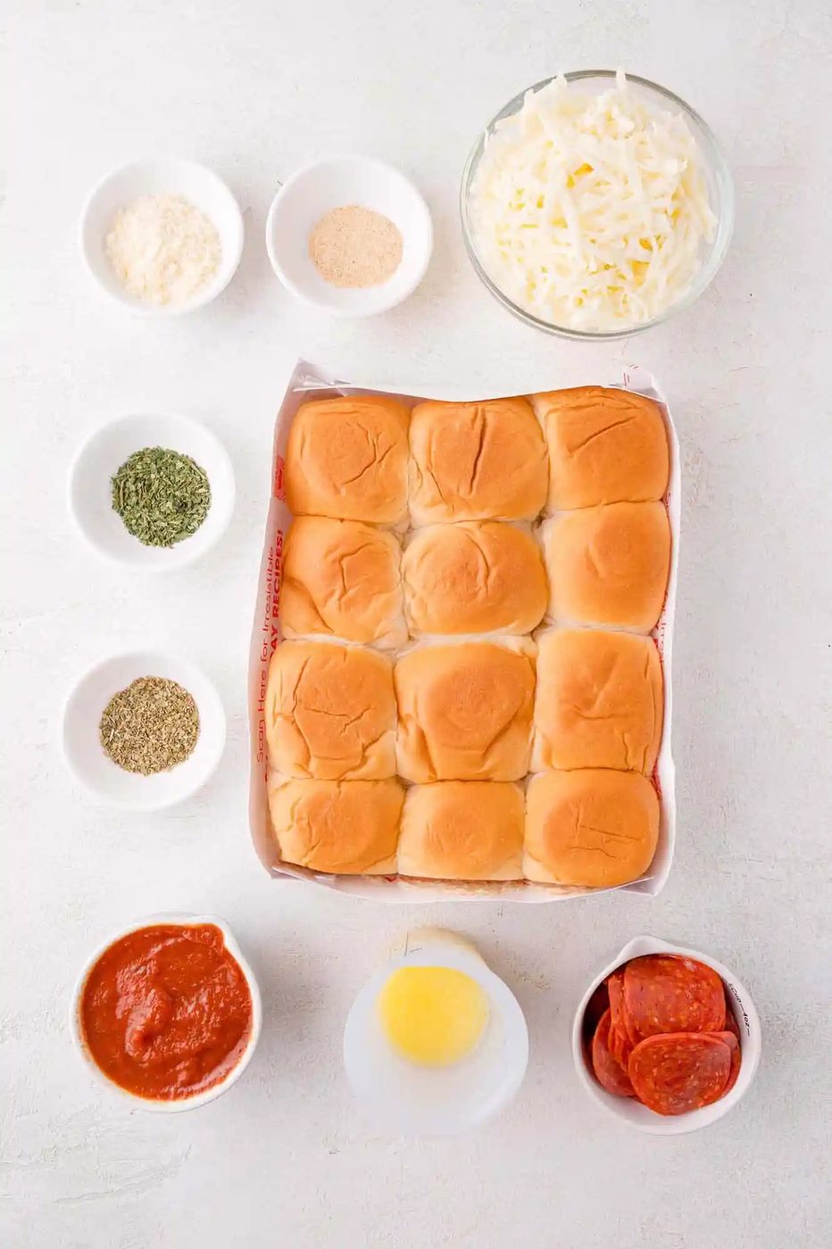 Top-down view of slider buns in a tray surrounded by bowls of shredded cheese, pepperoni slices, marinara sauce, melted butter, and seasonings on a white surface.