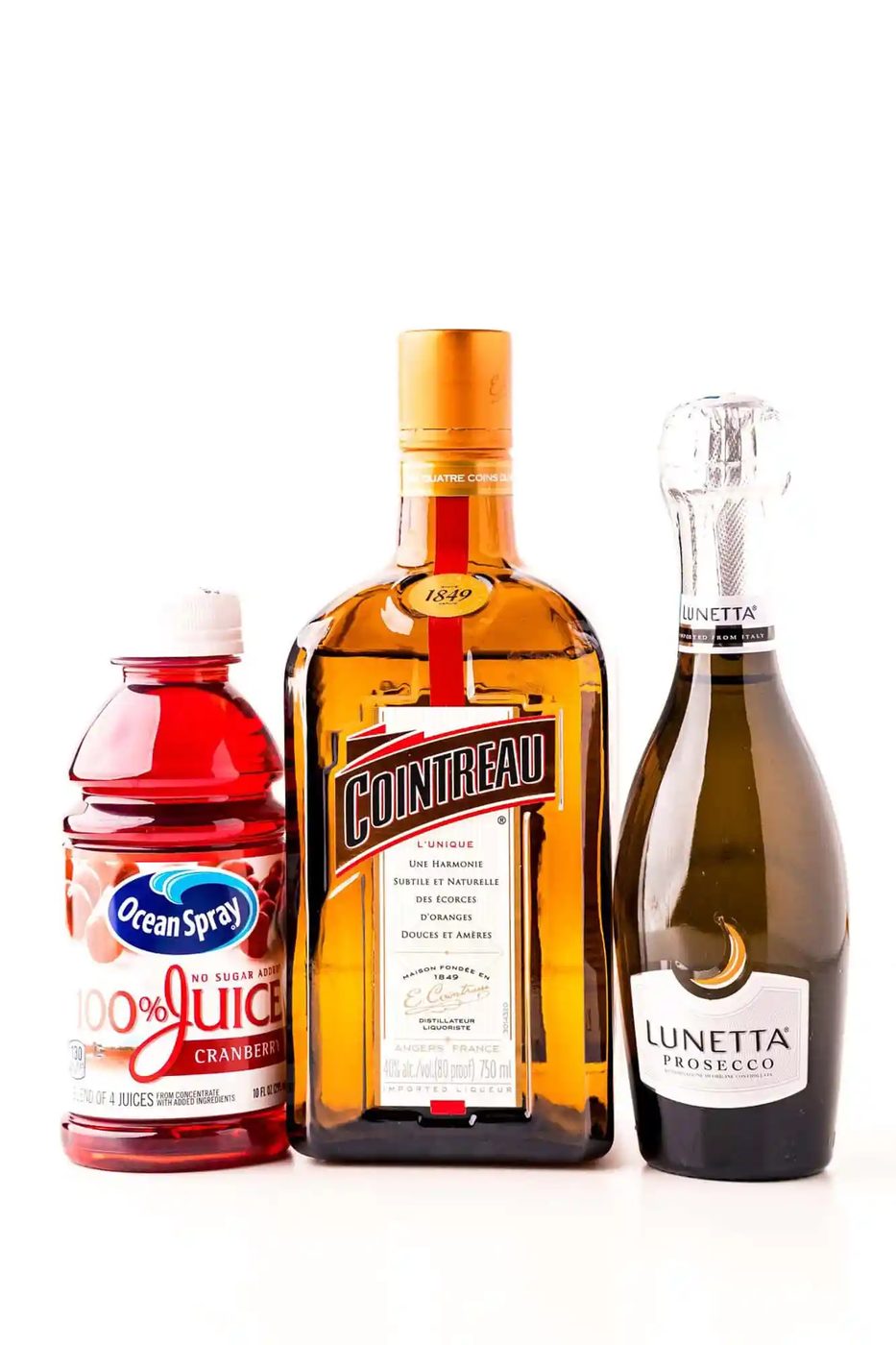 A bottle of cranberry juice, a bottle of Cointreau liqueur, and a bottle of Lunetta Prosecco are shown side by side against a white background.