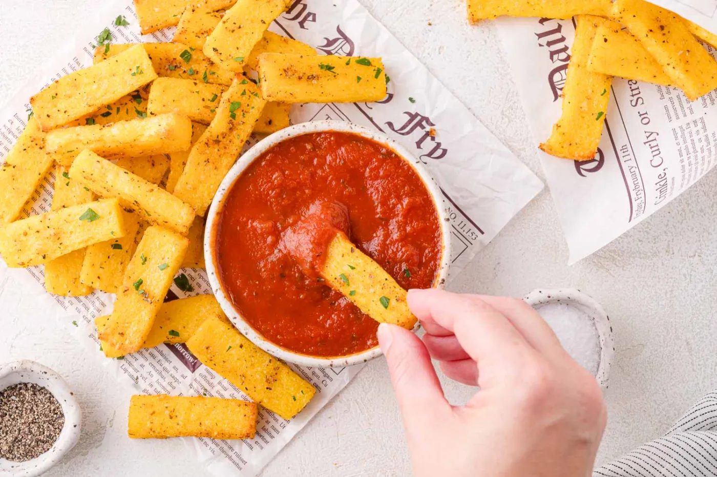 A hand dips a seasoned polenta fry into a bowl of red marinara sauce, with more fries and a small dish of pepper on the side.