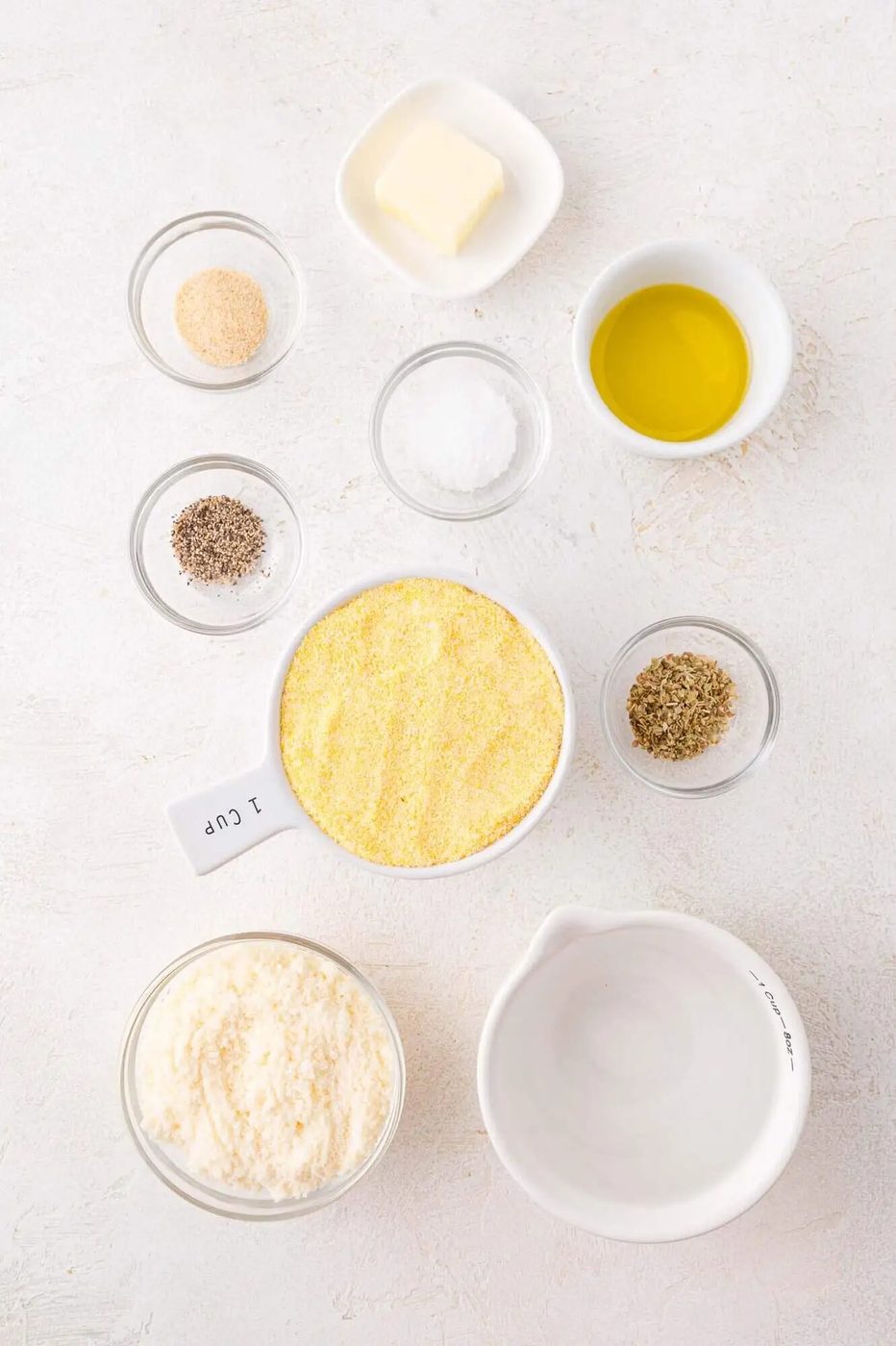 Bowls containing cornmeal, grated cheese, olive oil, butter, water, salt, pepper, oregano, and garlic powder arranged on a white surface.