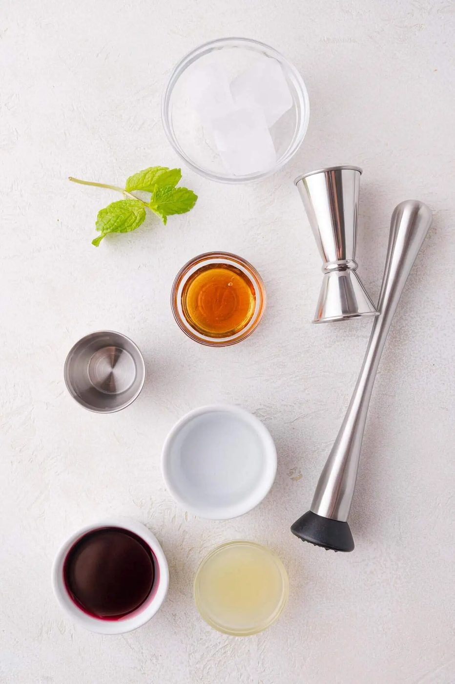 Assorted cocktail ingredients including ice cubes, fresh mint, lime juice, pomegranate juice, agave, and a metal jigger and muddler arranged on a white surface.