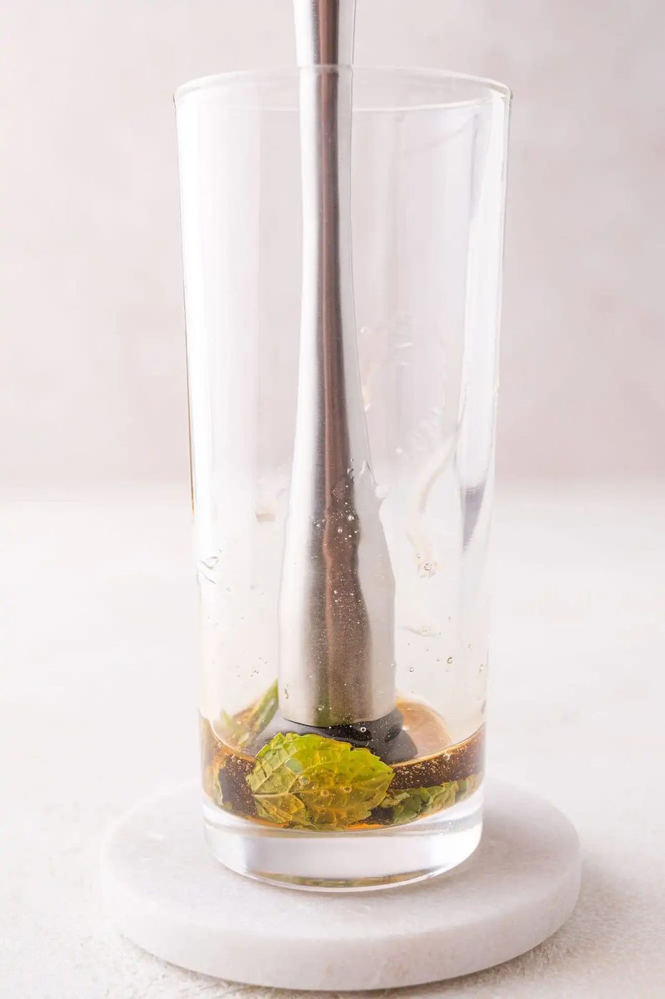 A metal muddler pressing mint leaves and agave at the bottom of a clear glass on a white coaster.