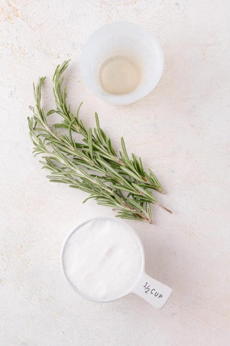 A plastic cup with water, a sprig of fresh rosemary, and a white measuring cup filled with sugar on a light surface.