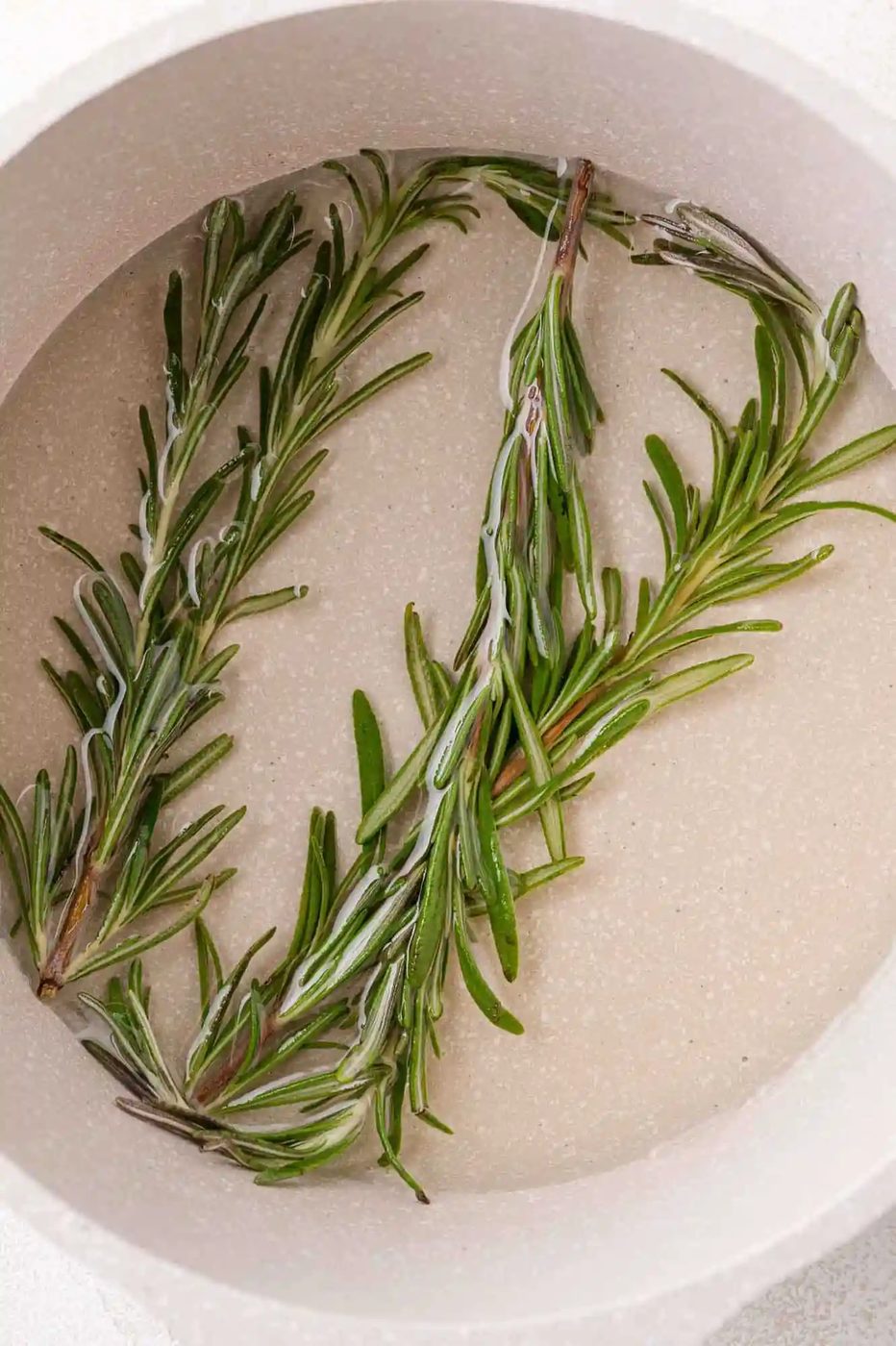 Two sprigs of fresh rosemary submerged in simple syrup inside a light-colored pot.