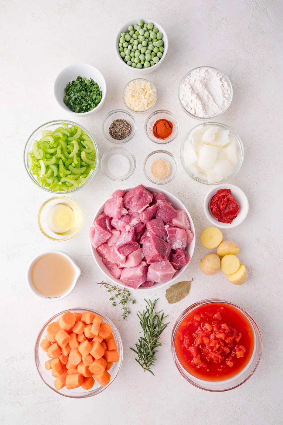 Assorted stew ingredients including raw pork, diced tomatoes, carrots, celery, peas, potatoes, onions, spices, herbs, tomato paste, oil, and flour arranged in bowls on a white surface.