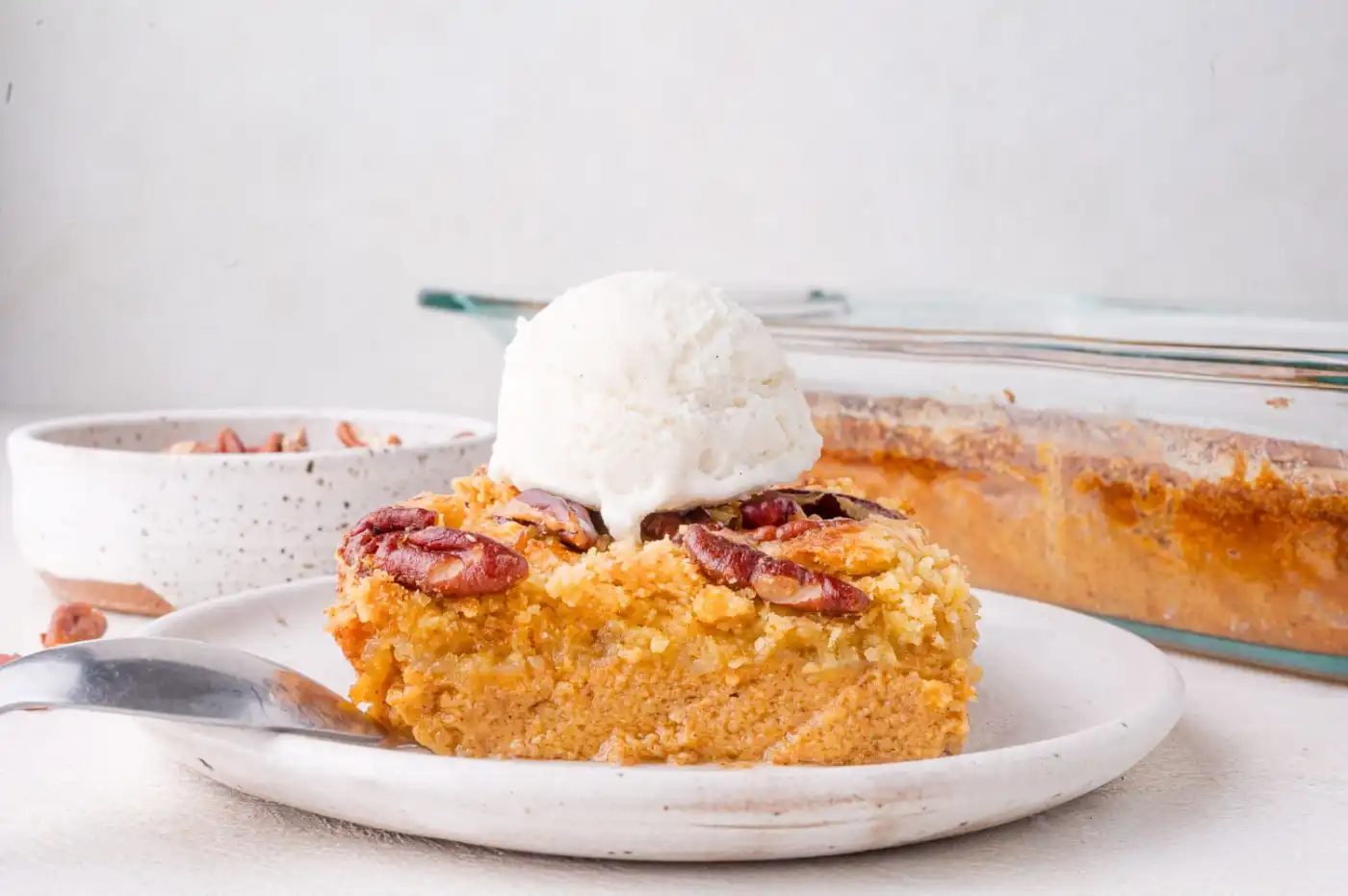 A slice of pumpkin dessert topped with pecans and a scoop of vanilla ice cream on a white plate, with a fork and a baking dish in the background.