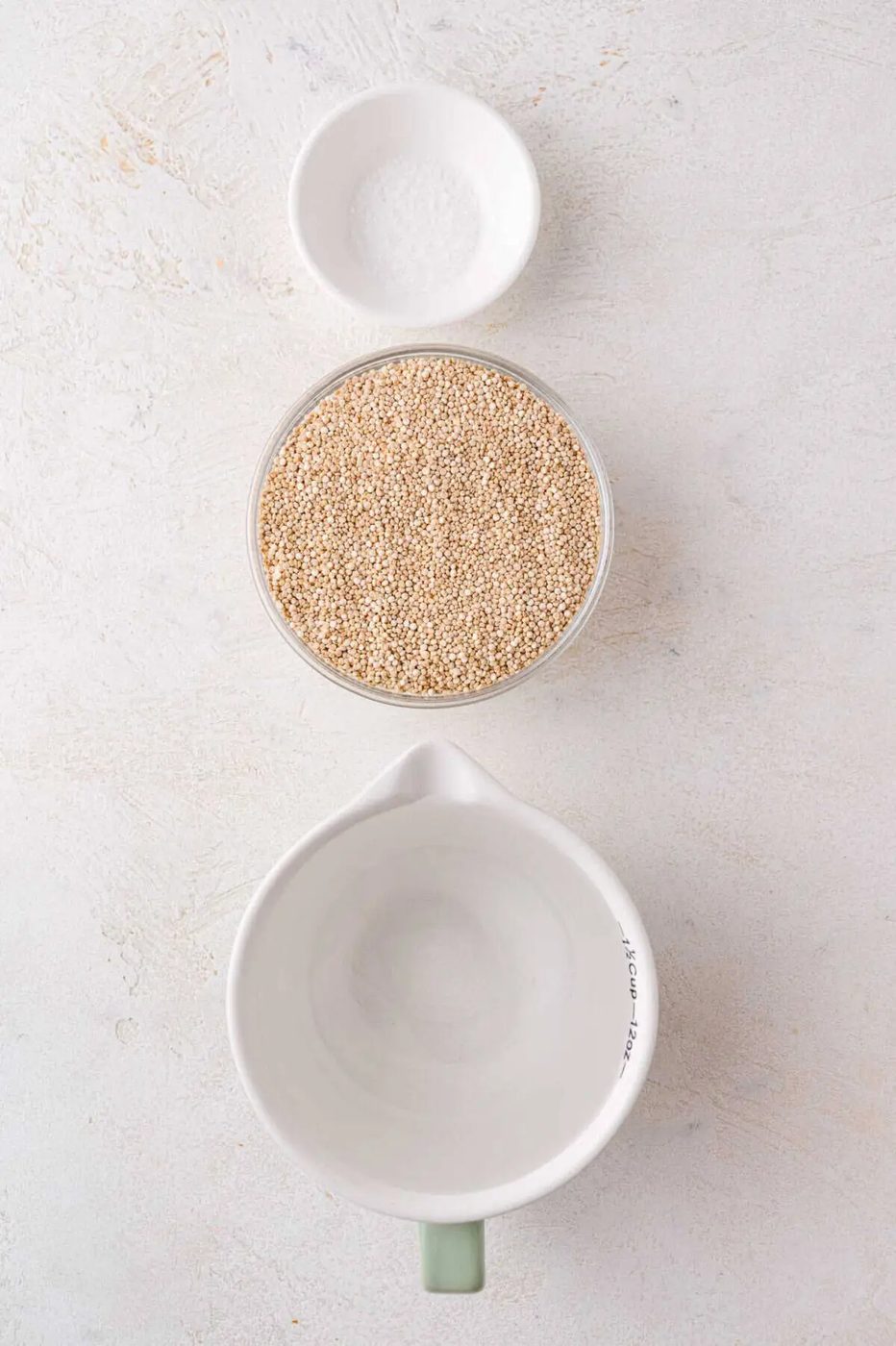 A small bowl of salt, a bowl of uncooked quinoa, and an empty measuring cup are arranged vertically on a light surface.