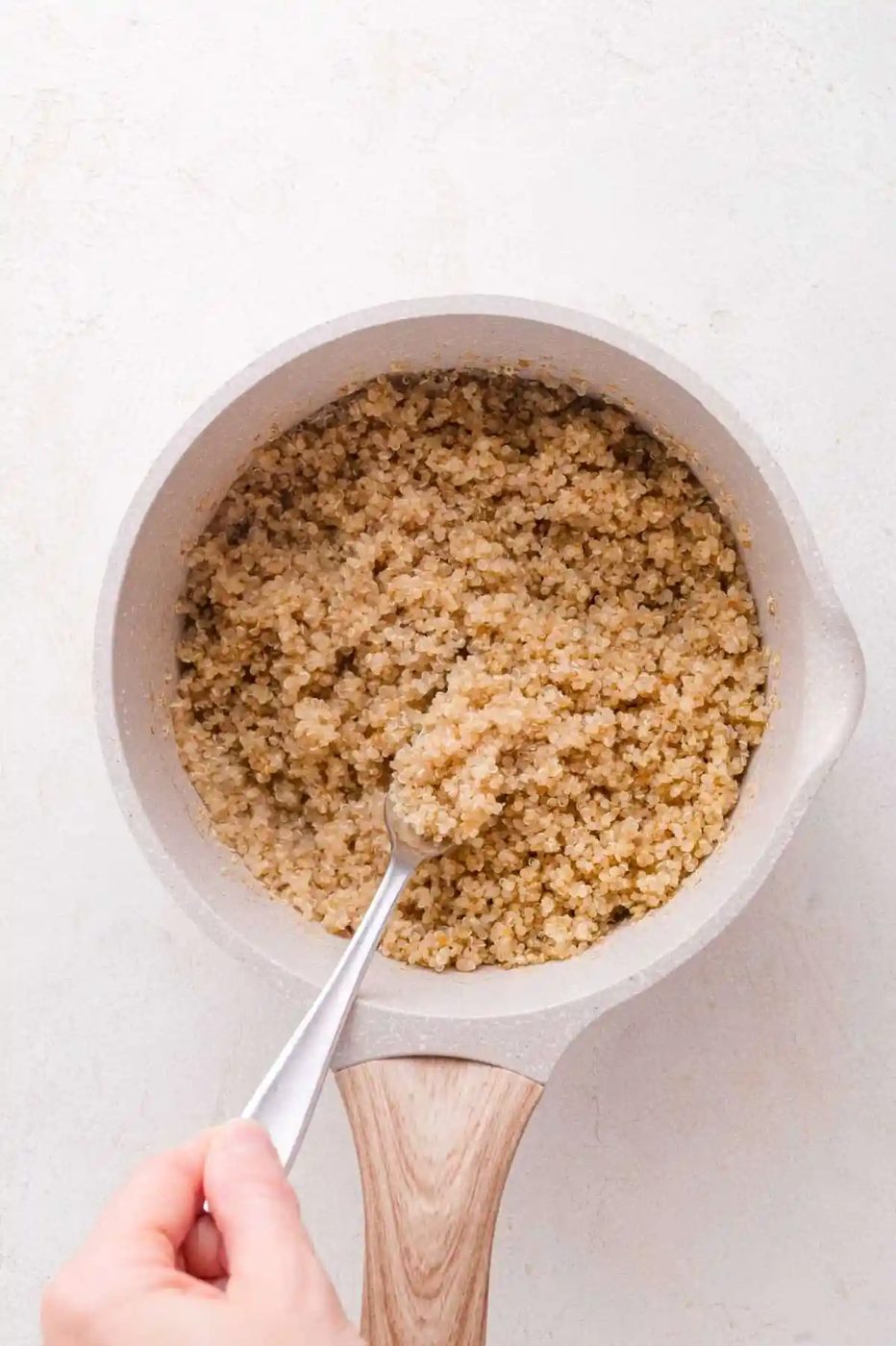 A hand holding a fork fluffs cooked quinoa in a light-colored saucepan with a wooden handle on a white surface.