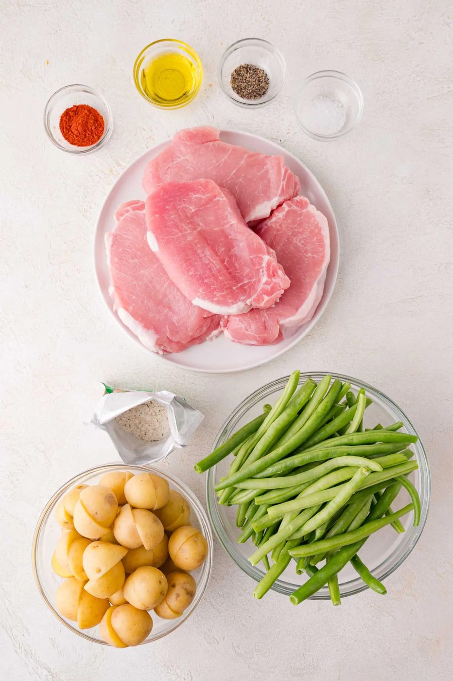 Raw pork chops on a plate with green beans, baby potatoes, and small bowls of paprika, olive oil, salt, and pepper arranged on a white surface.