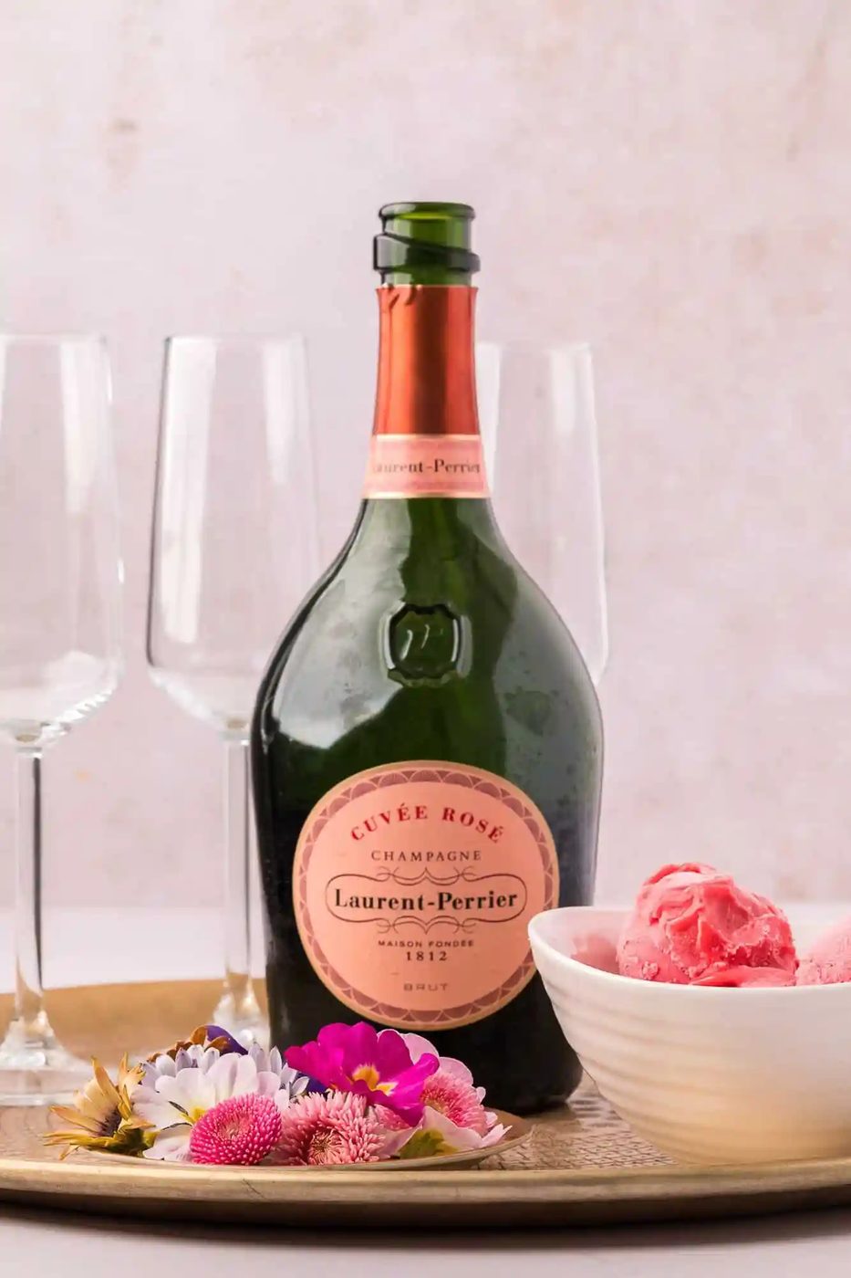 A bottle of Laurent-Perrier Cuvée Rosé champagne on a tray with four empty champagne flutes, a bowl of pink sorbet, and some colorful flowers.
