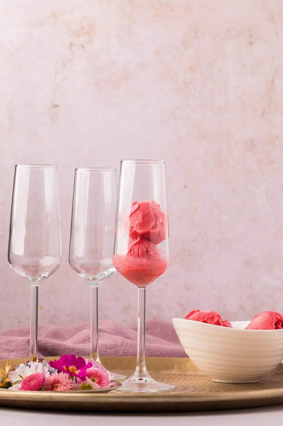 Three champagne flutes on a tray; one contains scoops of pink sorbet. A white bowl with more sorbet and some flowers are also on the tray, against a light pink background.