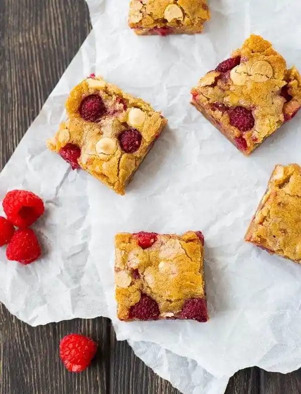 Blondie squares arranged on parchment paper with fresh raspberries scattered around.