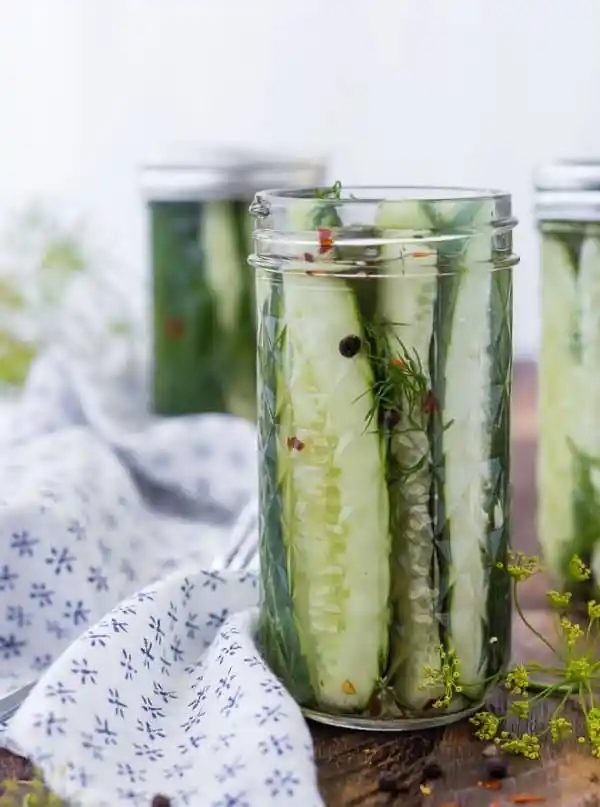 Image of from-scratch refrigerator dill pickles in a jar. Two jars in the background. Sprigs of dill also pictured.